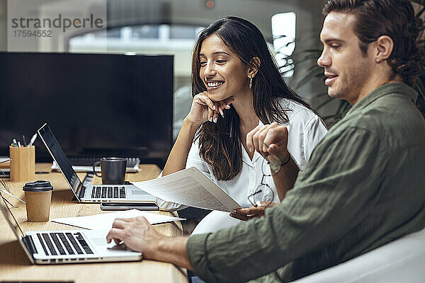 Lächelnder Geschäftsmann und Geschäftsfrau diskutieren am Schreibtisch im Coworking-Büro über Dokumente und Laptop