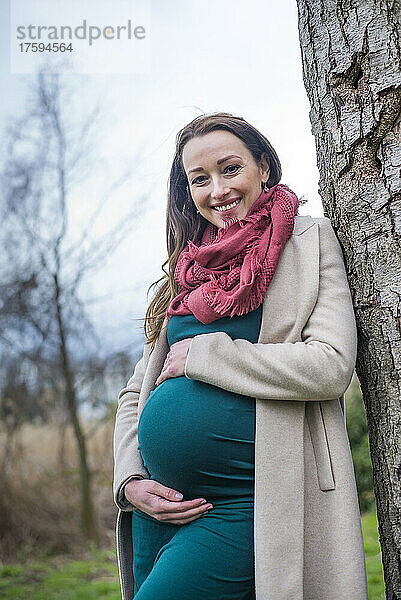 Smiling pregnant woman leaning on tree trunk