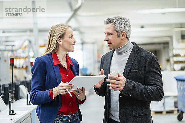Lächelnde blonde Geschäftsfrau mit Tablet-PC blickt auf Kollegen  der in der Fabrik Ideen bespricht