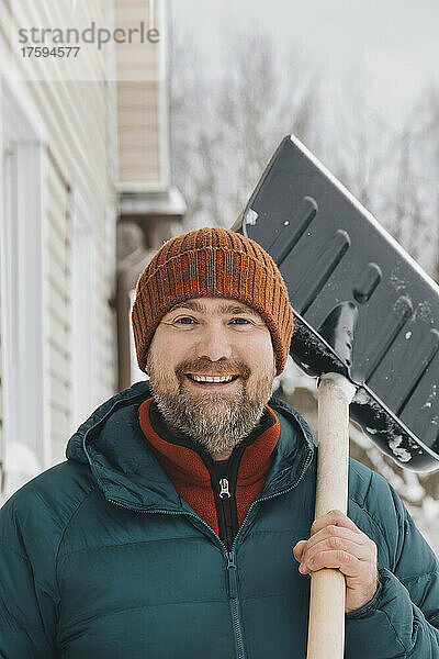 Glücklicher Mann mit Strickmütze und Schneeschaufel auf der Schulter im Winter