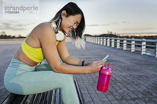 Young woman with smart phone and water bottle sitting on bench
