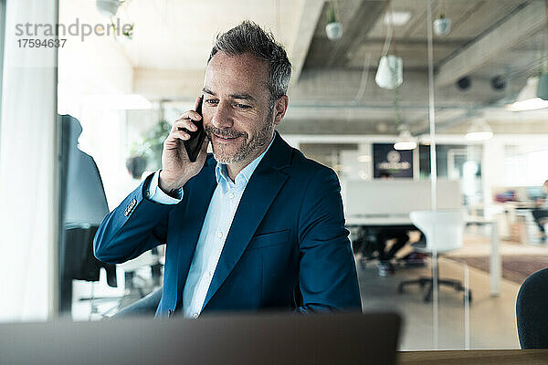 Smiling businessman talking on mobile phone looking at laptop in workplace