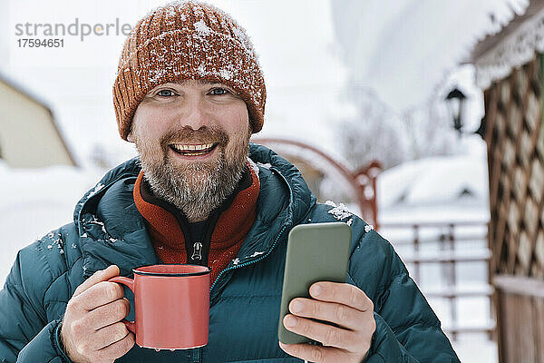 Lächelnder Mann mit Kaffeetasse und Smartphone im Winter