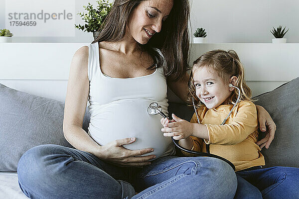 Lächelnde Tochter mit Stethoskop von schwangerer Mutter zu Hause im Bett
