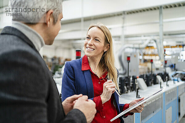 Lächelnde blonde Geschäftsfrau mit Tablet-PC blickt auf Kollegen  der in der Fabrik diskutiert