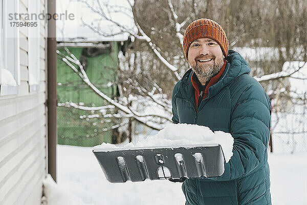 Lächelnder Mann hält Schneeschaufel mit Schnee im Winter