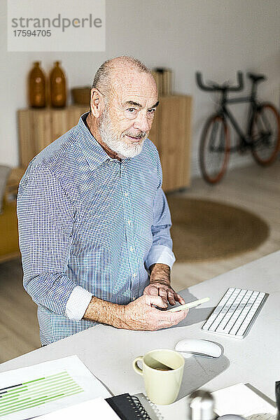 Nachdenklicher leitender Geschäftsmann mit Smartphone sitzt am Schreibtisch im Heimbüro