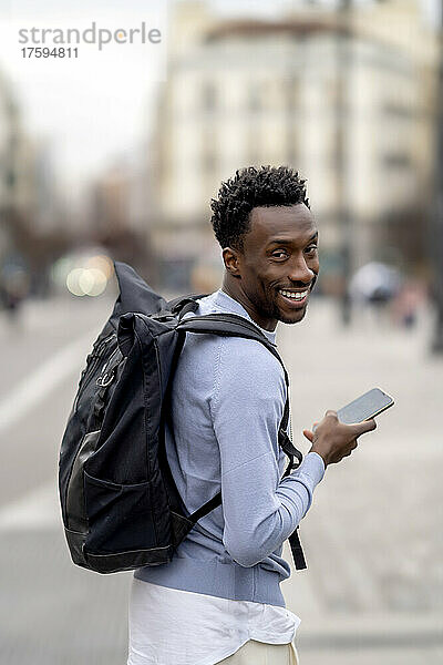 Lächelnder junger Mann mit Rucksack und Mobiltelefon