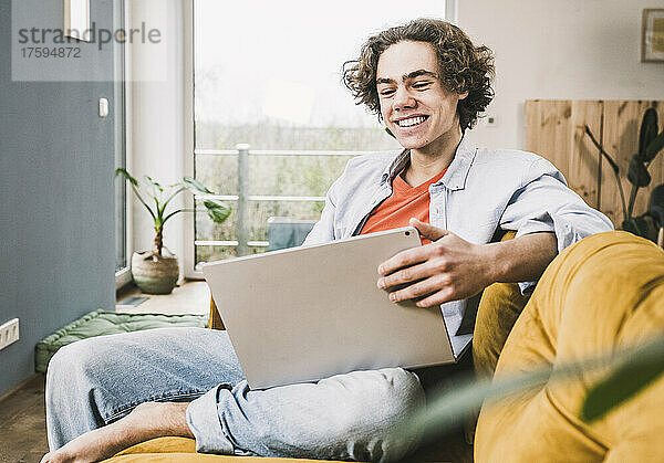 Glücklicher junger Mann sitzt mit Laptop auf dem Sofa im Wohnzimmer
