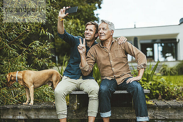 Älterer Mann gestikuliert mit Friedenszeichen und macht im Hinterhof ein Selfie mit seinem Sohn über sein Mobiltelefon