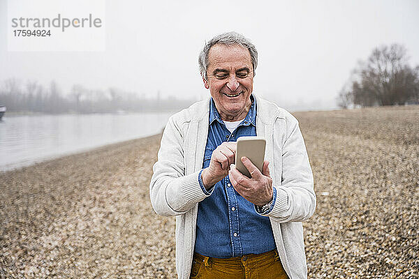 Glücklicher älterer Mann  der am Strand sein Mobiltelefon benutzt