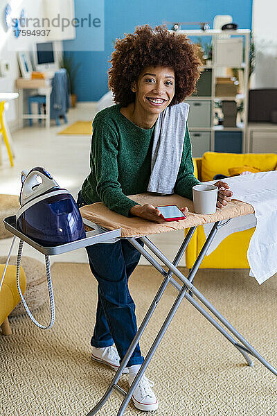Lächelnde Frau lehnt am Bügelbrett mit Kaffeetasse und Smartphone