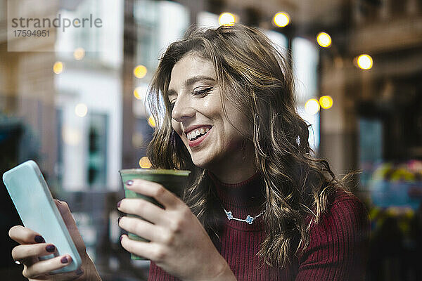 Lächelnde Frau mit Einwegkaffeetasse und Smartphone im Café