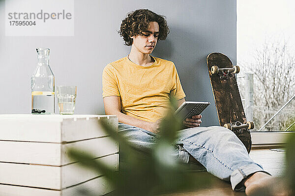 Junger Mann sitzt zu Hause am Skateboard und benutzt Tablet-PC