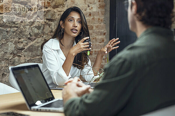 Geschäftsfrau hält Kaffeetasse in der Hand und diskutiert mit Geschäftsmann am Schreibtisch im Büro