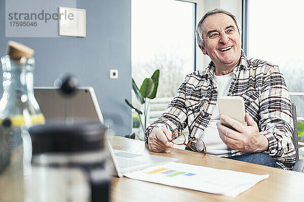 Fröhlicher älterer Mann mit Smartphone und Brille sitzt am Tisch
