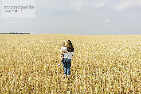 Mutter trägt ihren Sohn und geht an einem sonnigen Tag im Weizenfeld spazieren