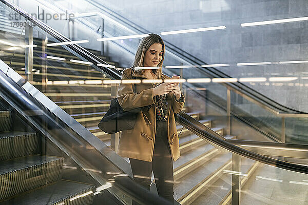 Lächelnde Frau benutzt Smartphone auf Rolltreppe einer U-Bahn-Station