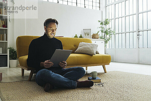 Reifer Mann sitzt zu Hause mit Tablet-PC auf dem Teppich