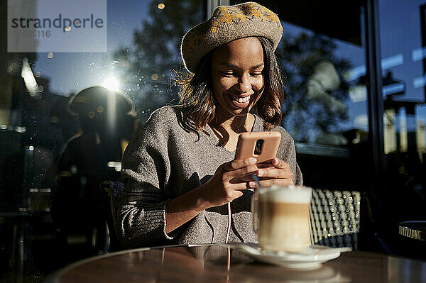 Lächelnde Frau mit Baskenmütze fotografiert Kaffeetasse auf dem Tisch im Café