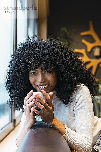 Smiling curly black haired woman with coffee cup leaning on sofa at cafe