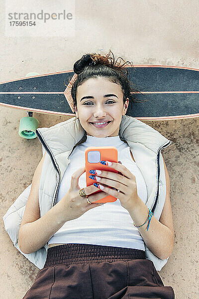 Junge Frau mit Smartphone liegt auf Skateboard im Park
