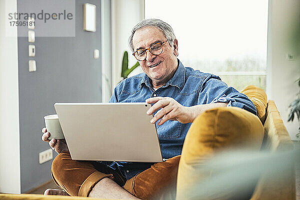 Älterer Mann hält Kaffeetasse in der Hand und schaut sich im heimischen Wohnzimmer ein Video auf dem Laptop an