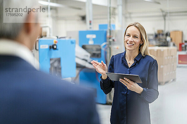 Lächelnde blonde Geschäftsfrau hält Tablet-PC in der Hand und diskutiert mit Kollegen in der Fabrik