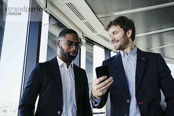 Lächelnder Geschäftsmann mit Smartphone blickt Kollegen im Büro an