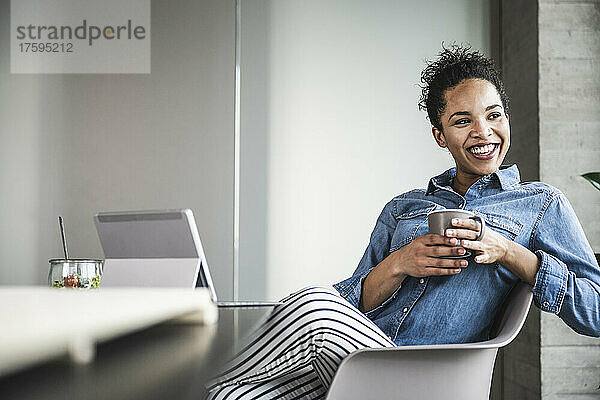 Glückliche Geschäftsfrau mit Kaffeetasse sitzt auf einem Stuhl im Büro