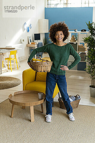 Lächelnde Afro-Frau mit der Hand an der Hüfte und einem Korb im Wohnzimmer