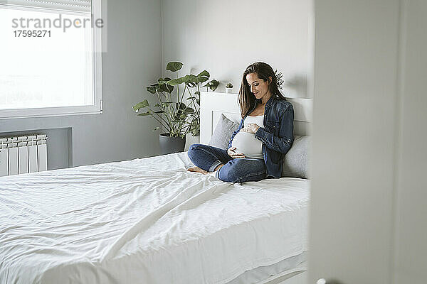 Schwangere Frau sitzt mit gekreuzten Beinen und berührt den Bauch zu Hause im Bett
