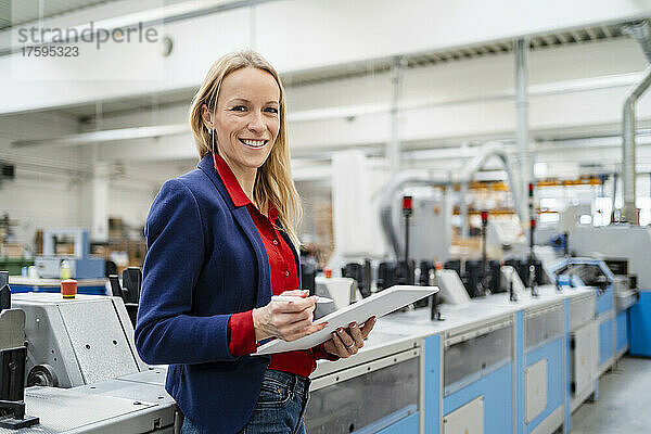 Glückliche blonde Geschäftsfrau mit Tablet-PC  die neben Maschinen in der Industrie steht