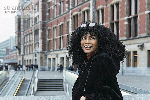Lächelnde junge Frau mit lockigem Haar  die in der Stadt über die Schulter schaut