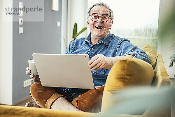 Fröhlicher älterer Mann mit Laptop und Kaffeetasse sitzt auf dem Sofa im Wohnzimmer
