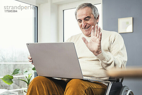 Lächelnder behinderter Mann bei Videoanruf über Laptop winkt mit der Hand im Rollstuhl