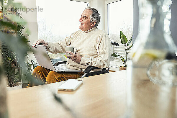 Glücklicher älterer behinderter Mann mit Laptop und Kaffeetasse  der zu Hause im Rollstuhl sitzt