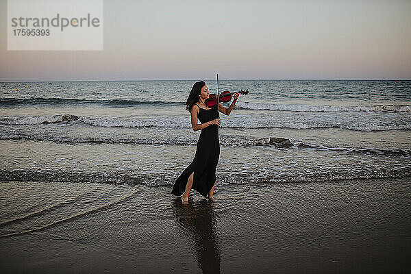 Frau spielt Geige und steht bei Sonnenuntergang vor dem Meer