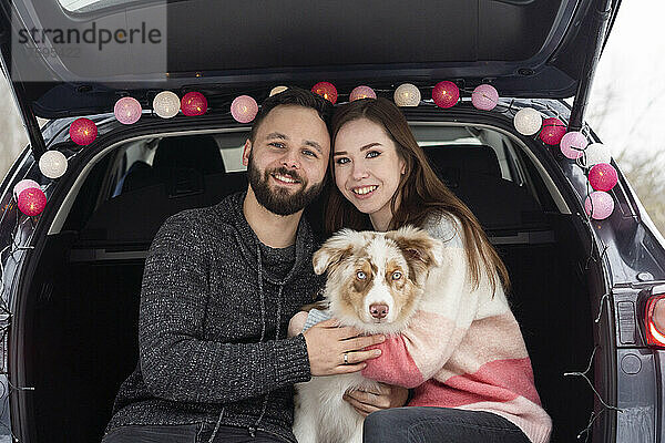 Glückliches junges Paar sitzt mit Hund im Kofferraum eines Autos