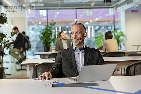 Lächelnder Geschäftsmann mit Laptop am Schreibtisch im Coworking Space