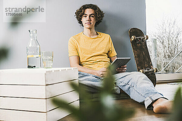 Junger Mann mit Tablet-PC sitzt zu Hause am Skateboard