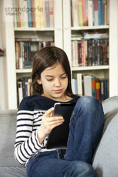 Junges Mädchen sitzt auf dem Sofa im Wohnzimmer mit digitalem Tablet in der Hand