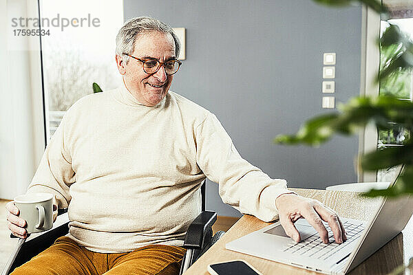 Lächelnder behinderter Mann mit Kaffeetasse und Laptop  der im Rollstuhl am Tisch sitzt