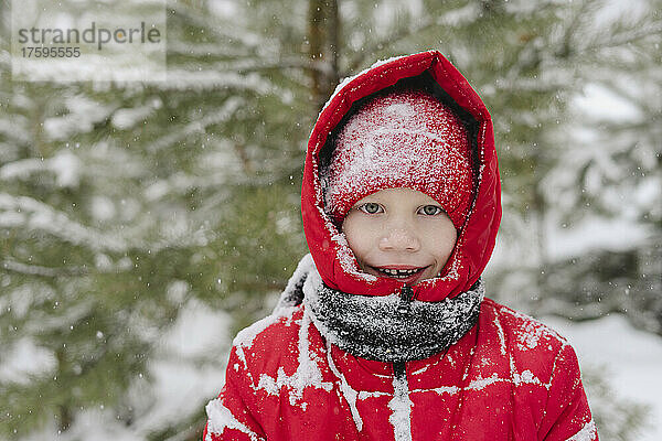 Fröhlicher Junge mit Schnee auf Strickmütze und warmer Kleidung im Wald