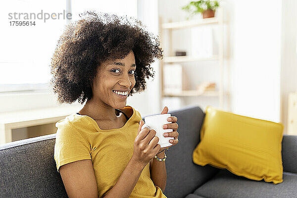 Glückliche junge Frau mit Kaffeetasse sitzt auf dem Sofa im Wohnzimmer