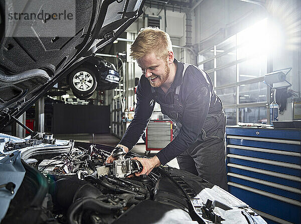 Lächelnder junger blonder Mechaniker repariert Automotor in Autowerkstatt