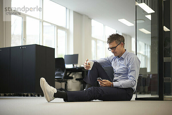 Geschäftsmann sitzt mit Smartphone auf dem Boden im modernen Büro