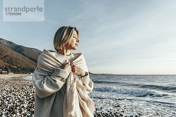 Blonde Frau  in eine Decke gehüllt  genießt den Sonnenuntergang am Strand