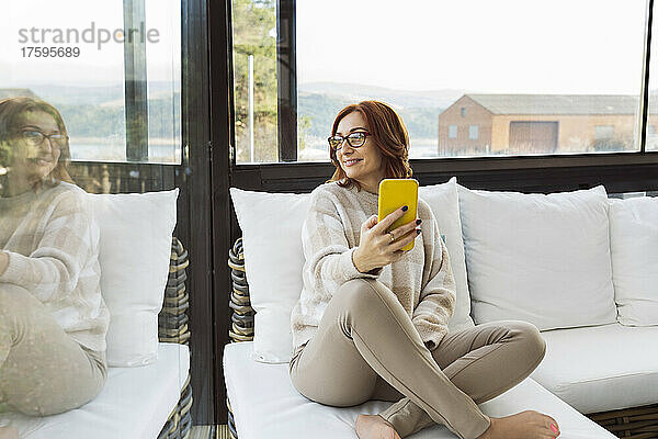 Lächelnde Frau mit Smartphone  die zu Hause durch das Fenster schaut
