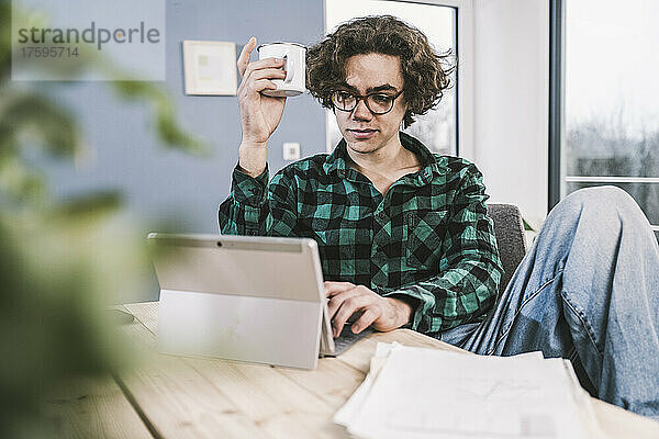 Mann hält Kaffeetasse mit Tablet-PC am Tisch im Wohnzimmer